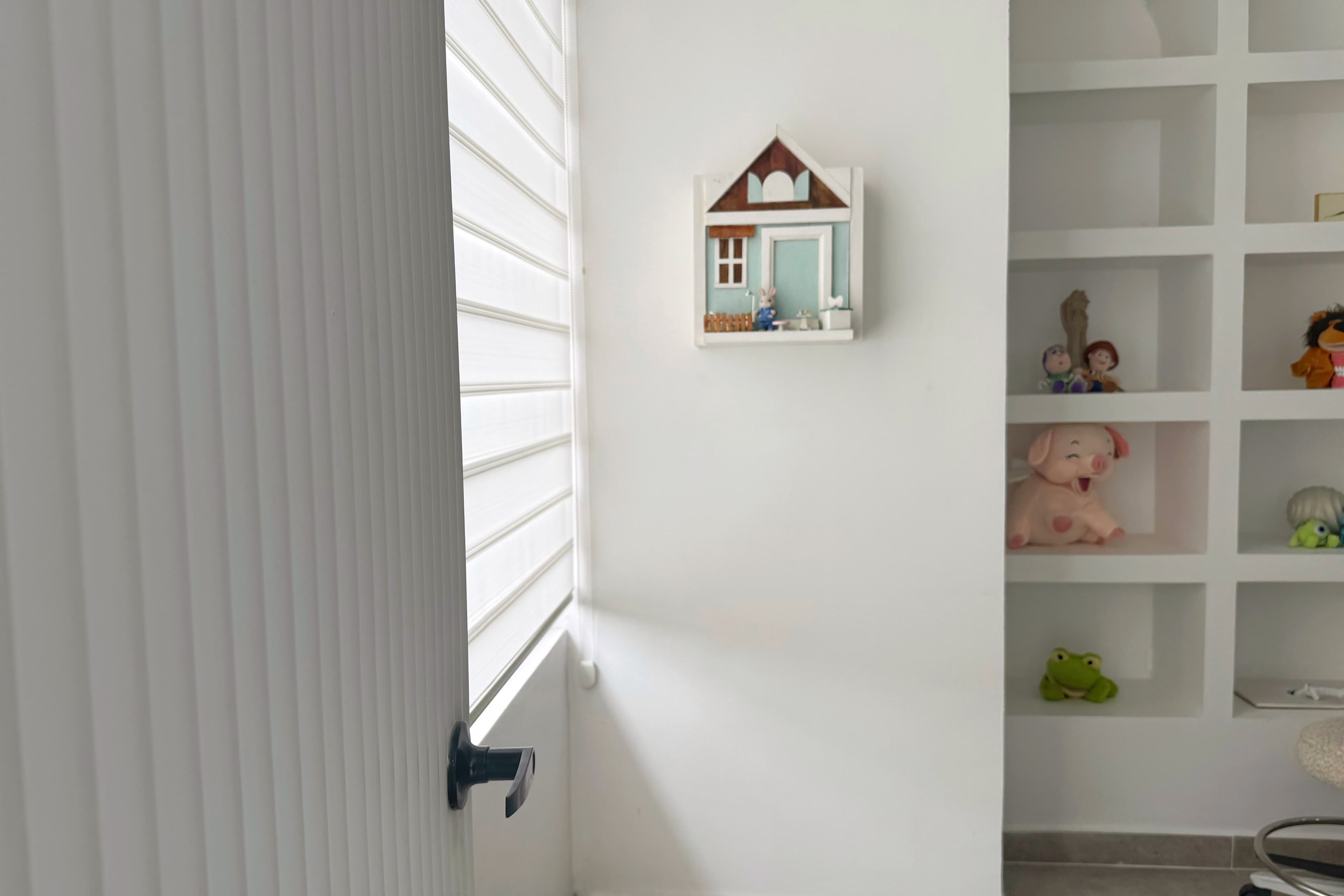 Detalle de consultorio odontopediátrico con estanterías blancas, juguetes decorativos, puerta acanalada y luz natural filtrada por persianas.