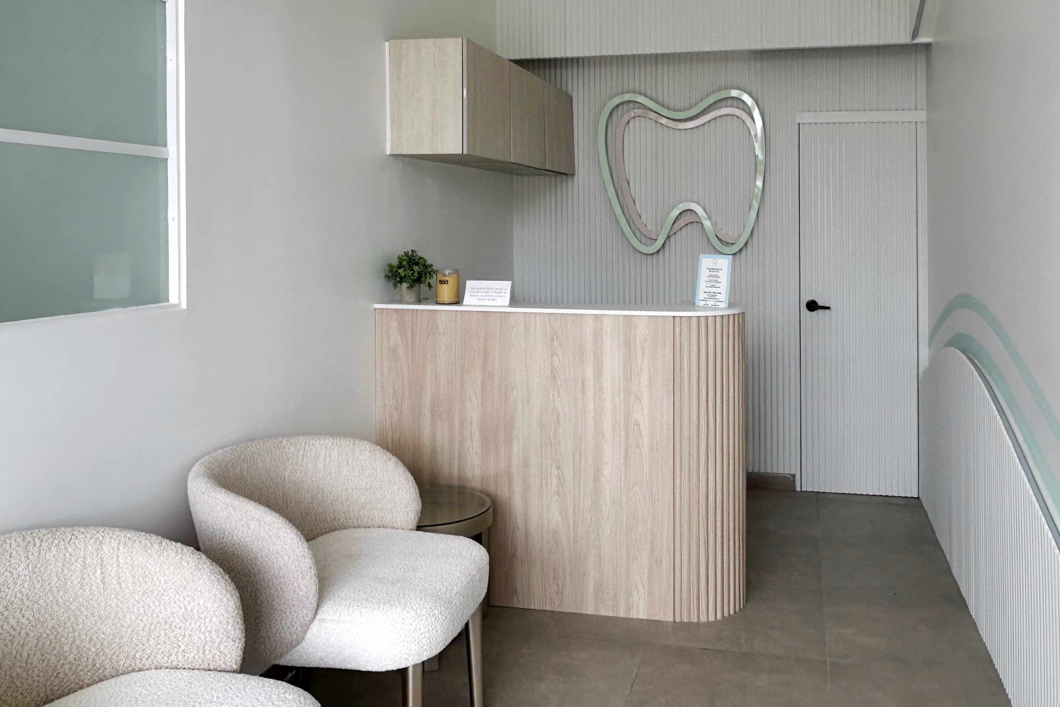 Área de recepción de clínica dental con mostrador curvo de madera clara, revestimientos acanalados, sillones blancos y detalles decorativos en tonos menta.