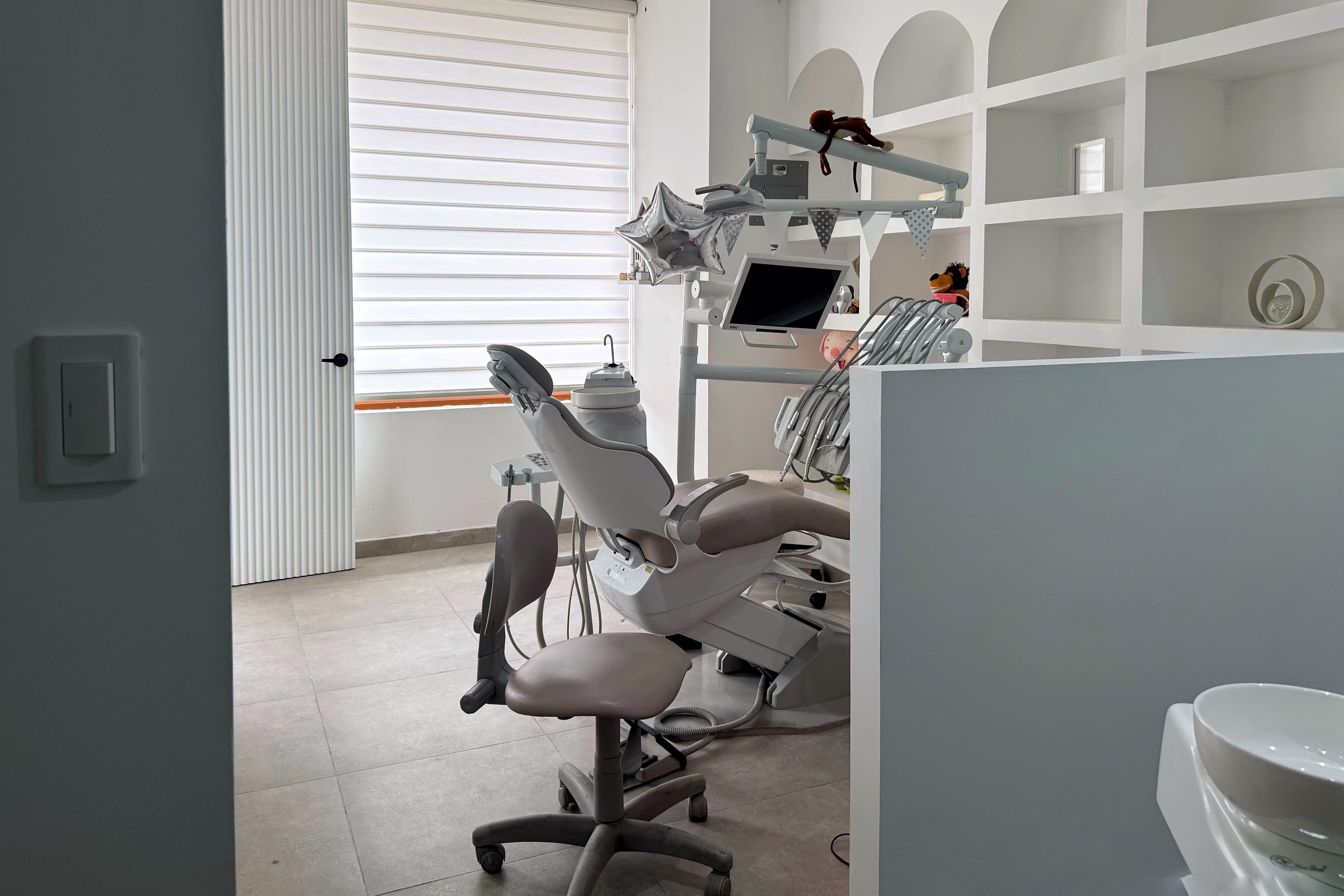 Interior de consultorio odontológico con sillón dental gris, equipamiento médico, mobiliario blanco de nichos arqueados e iluminación natural.